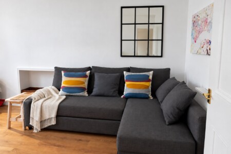 Grey corner sofa with colourful cushions and a cream throw beneath a black grid mirror, with artwork on the adjoining wall.