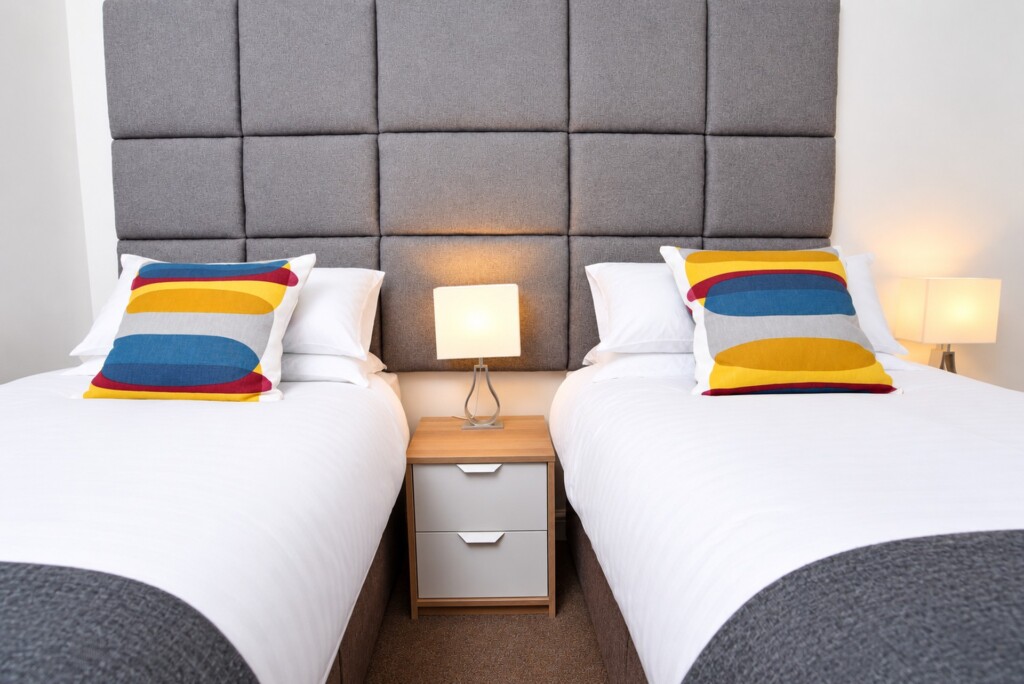 Two separate single beds with white bedding, grey throws and striped cushions, with a bedside cabinet between them.