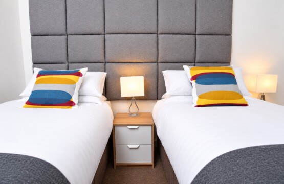 Two separate single beds with white bedding, grey throws and striped cushions, with a bedside cabinet between them.