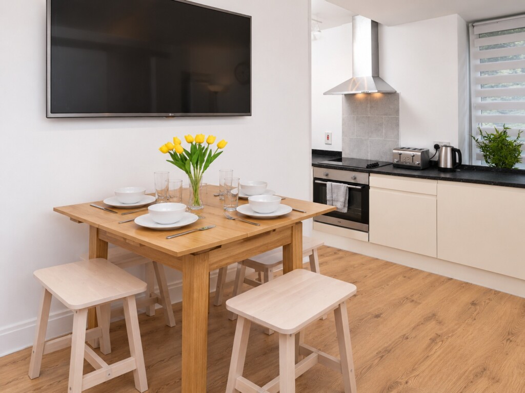 Wooden dining table and four pale stools beneath a wall-mounted television, beside white kitchen cabinets on a wood-effect floor.
