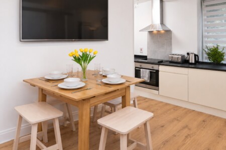 Wooden dining table and four pale stools beneath a wall-mounted television, beside white kitchen cabinets on a wood-effect floor.