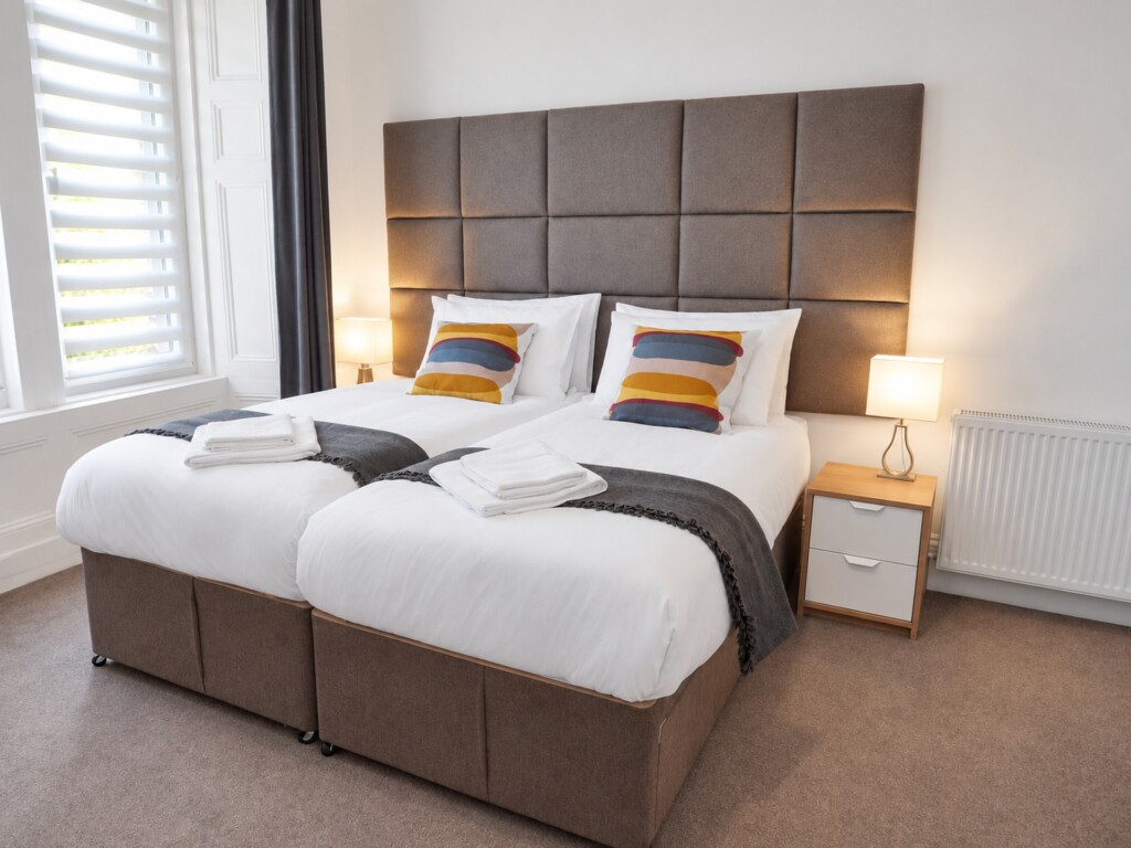 Beds arranged together beneath a tall grey upholstered headboard, beside a window with a blind and bedside lighting.
