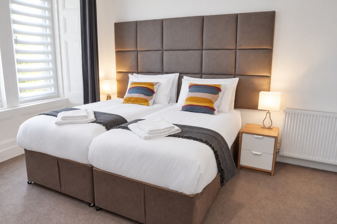 Beds arranged together beneath a tall grey upholstered headboard, beside a window with a blind and bedside lighting.