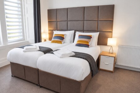 Beds arranged together beneath a tall grey upholstered headboard, beside a window with a blind and bedside lighting.