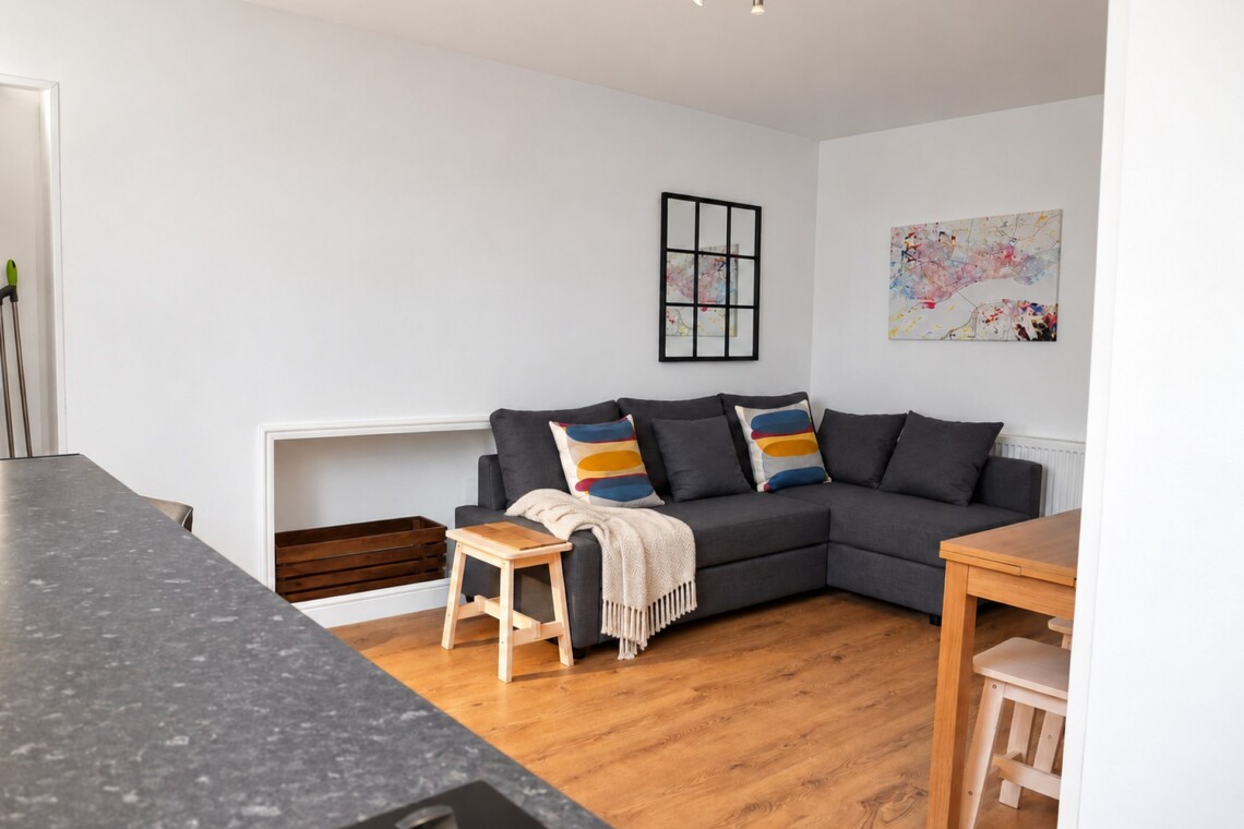 View across a kitchen worktop towards a grey corner sofa, cream throw, black grid mirror and artwork, with a table to the right.