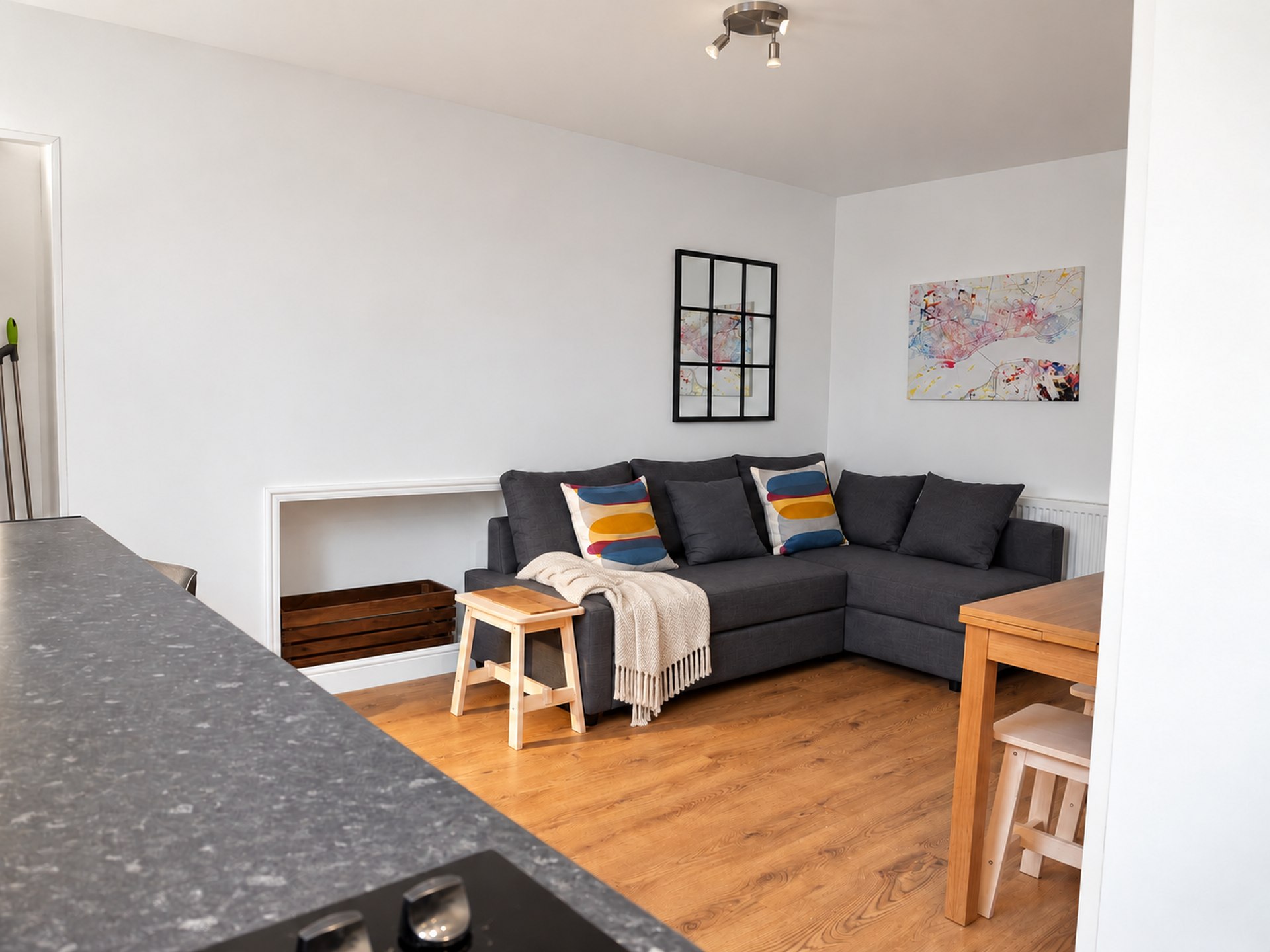 View across a kitchen worktop towards a grey corner sofa, cream throw, black grid mirror and artwork, with a table to the right.